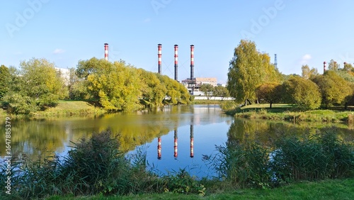 Trees grow on the grassy banks of the river and are reflected in the water. There are reeds in the water. On the opposite bank are city buildings and administrative buildings with factory chimneys