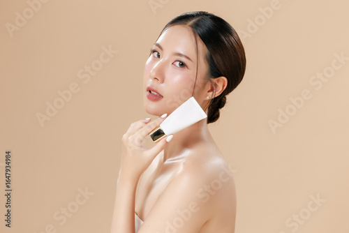 Young Asian woman portrait natural make up cosmetic on face radiant eyes lip gloss happy touch her skin on isolated beige studio. Female model Holding skincare product Facial concept.