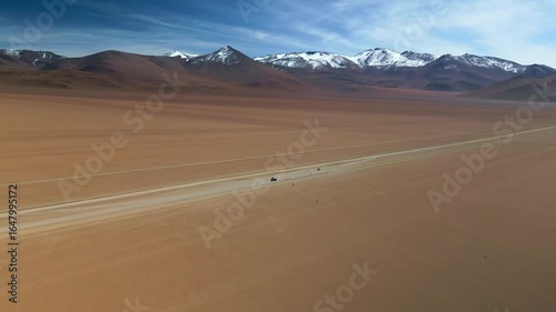 Drone flying forward over Dali Desert with camera tilting down, jeep parked on dirt road with people nearby, snow-capped mountains and blue sky background.