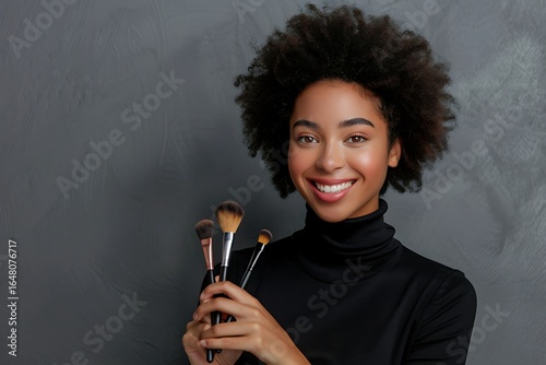 Black woman with afro hairstyle, Holding makeup brushes, Studio portrait on gray background, Confident beauty artist pose, Minimal backdrop, Copy space for text