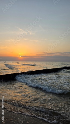 Gentle waves crash against the structure, creating white foam, while the low sun near the horizon bathes the sky in warm shades of orange, pink, and gold. In the background, the calsunset over the sea