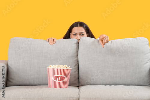 Young woman watching movie behind sofa on yellow background