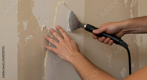 DIY wallpaper stripping: Close-up of hands using a steam scraper to remove old peeling wallpaper from a wall during home renovation.