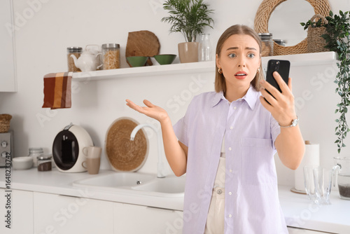 Upset young woman with mobile phone video chatting in kitchen