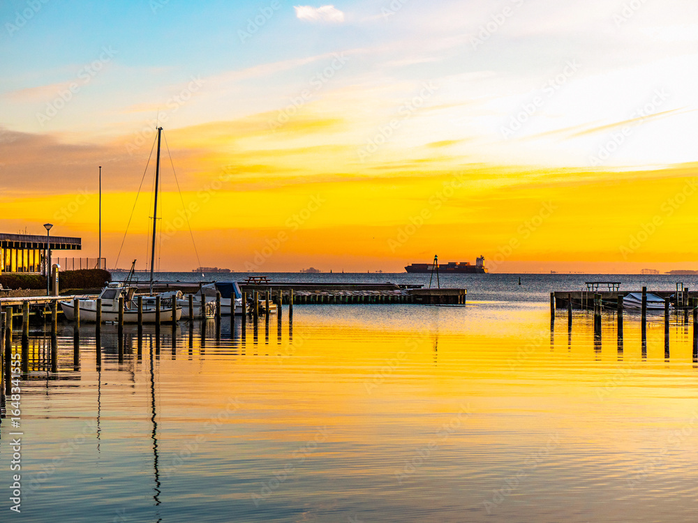 Obraz premium Sunrise over calm waters at Kastruphavn marina in Tårnby, Denmark, with boats, wooden piers, and a distant ship under a vibrant orange and blue sky.