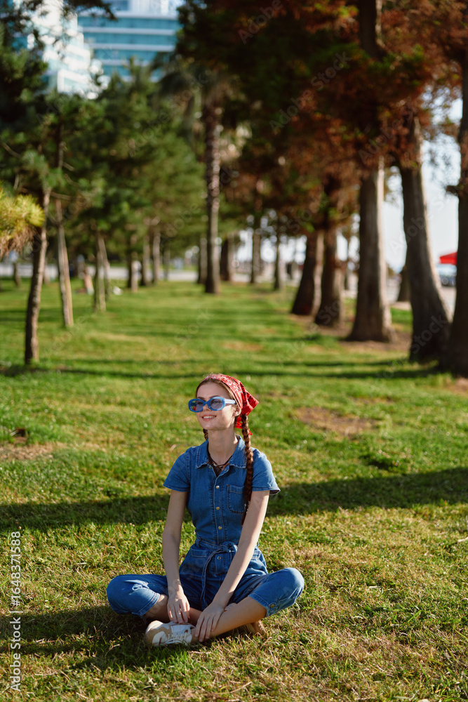 Fototapeta premium Lifestyle woman in modern boho-western denim street style sitting cross-legged on grass, wearing sunglasses and red headscarf, sunny outdoor park with trees and urban background.