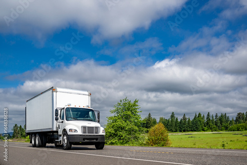 Compact middle duty day cab white rig semi truck transporting cargo in box trailer running on the local highway intersection