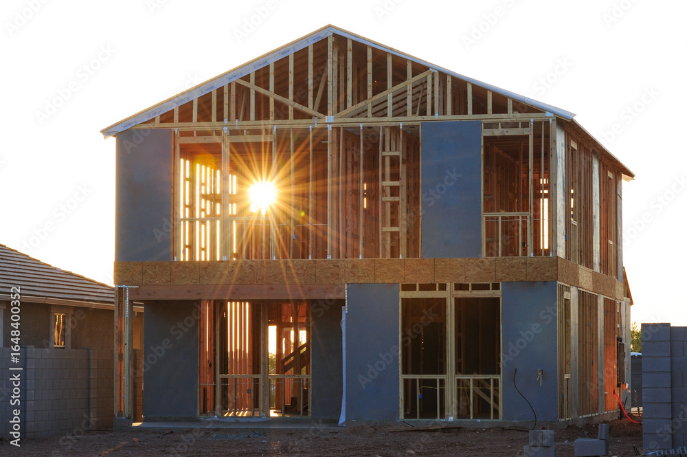 Obraz premium Two-story house under construction with exposed wooden framing and sheathing panels, captured at sunrise during the structural phase of residential building in a suburban housing development