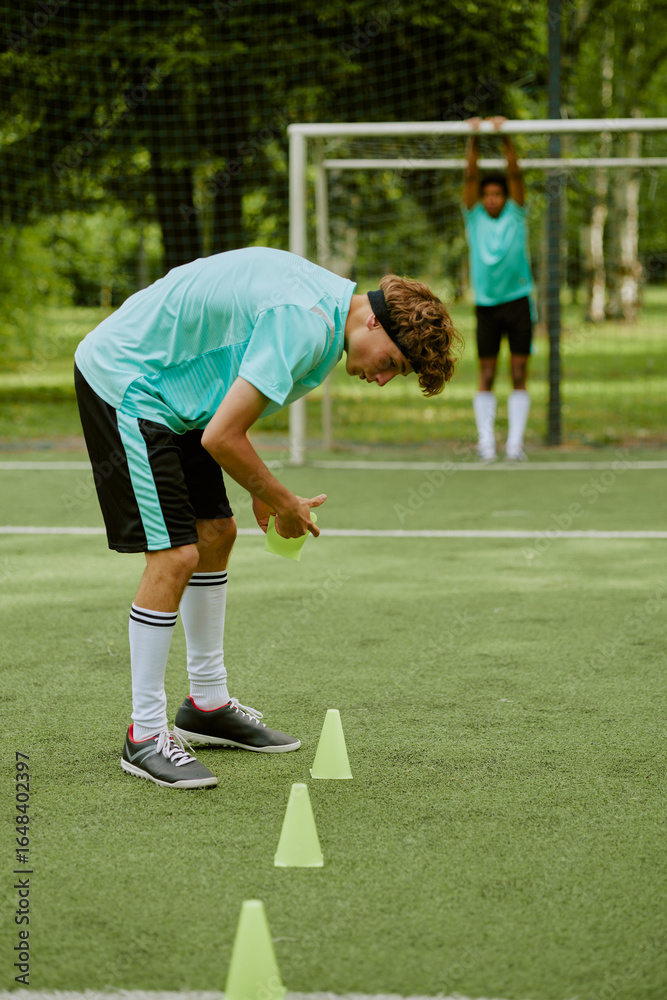 Obraz premium Teenage Caucasian boy arranging training cones on outdoor soccer field while Black teenage boy standing in background near goalpost stretching arms above head