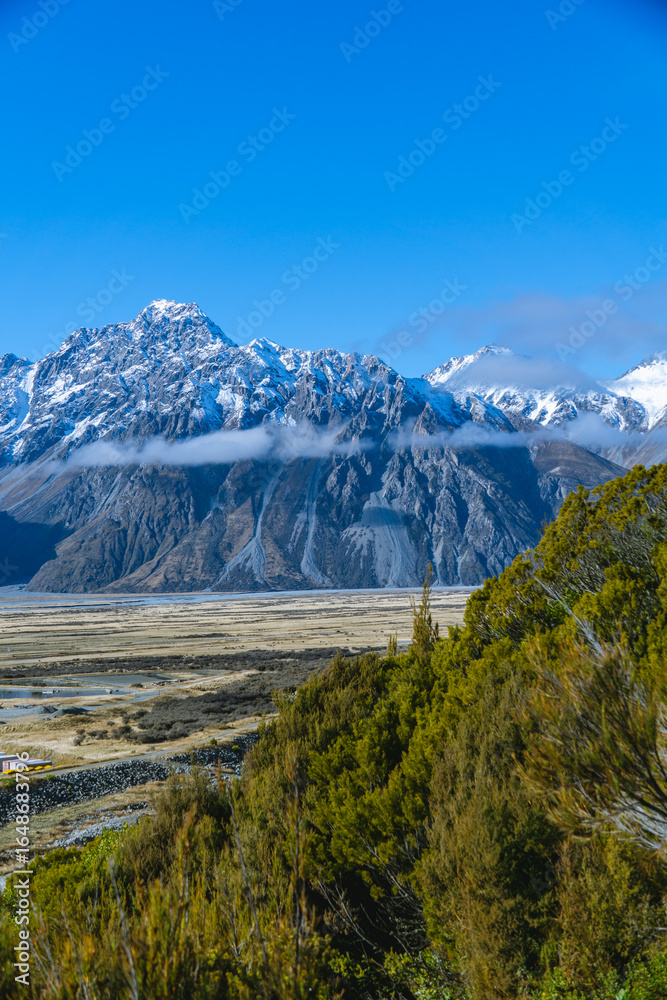 Obraz premium Majestic snow-covered peaks rise above a golden valley bordered by green forest.
