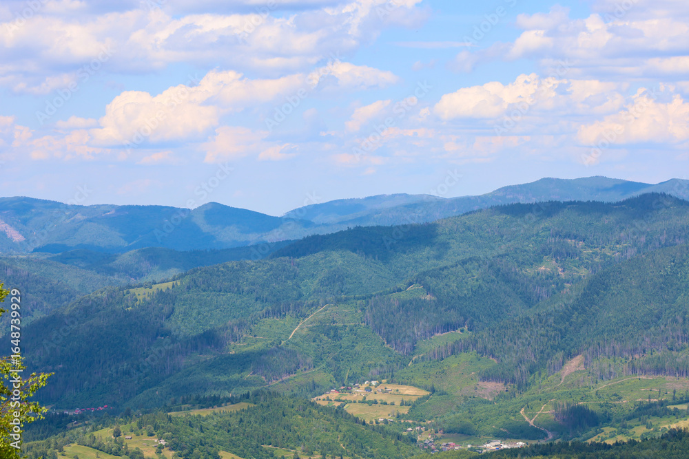Fototapeta premium Endless wooded mountain ranges stretching into the distance create a breathtaking panorama. Small villages are nestled among them, surrounded by greenery, and above this majestic landscape, a blue sky