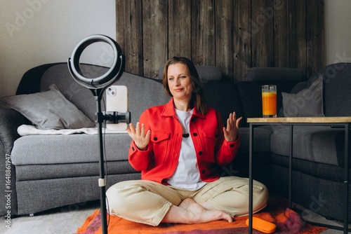 Smiling woman filming video with smartphone and ring light at home. Sitting cross-legged on the floor, she creates content, talks to camera, remote online communication or vlogging session