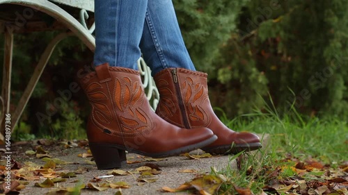 Close-up of legs in cowboy boots, beating time and rhythm to the music. Side view. Unrecognisable person sitting on bench in autumn park.