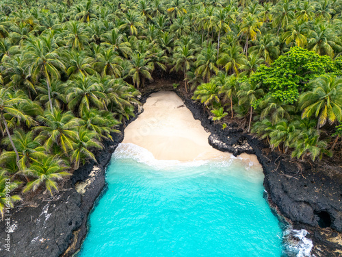 Tropical aerial view of Battery Beach on Rolas Island, São Tomé and Príncipe, showcasing turquoise water, black volcanic rocks, and dense green palm forest.
