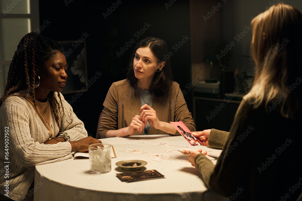 Fototapeta premium Three young women, Black and Caucasian, sitting around round table, engaged in mystic tarot reading session, discussing boyfriend on photograph, while consulting psychic for relationship advice