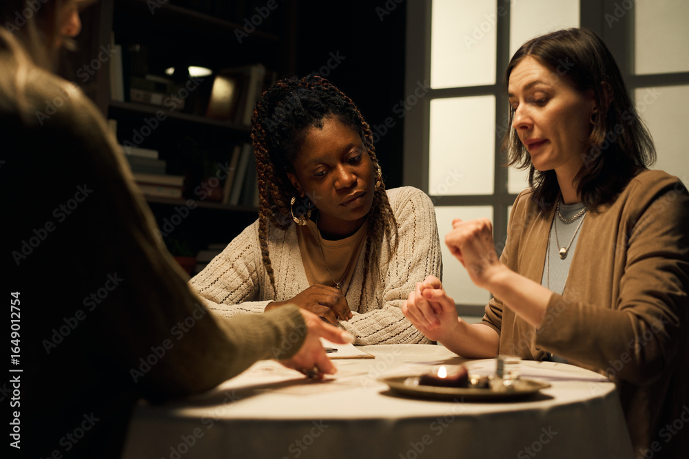 Fototapeta premium Caucasian young woman choosing tarot card on table during reading with female mystic, while African American girlfriend is watching, requesting love and relationship advice