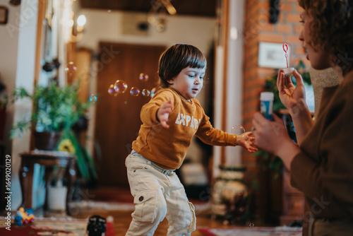 Fotografie A delightful indoor scene capturing a cheerful child chasing soap bubbles as their mother blows them