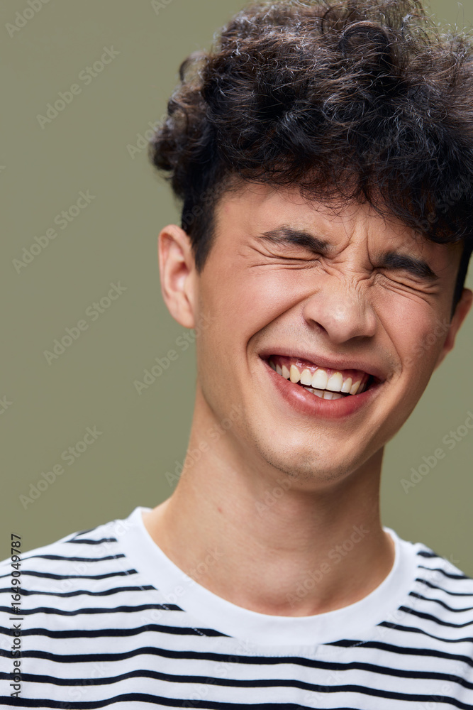 Fototapeta premium smiling man with curly hair in striped shirt laughing with closed eyes, natural joyful expression, studio portrait with muted green background