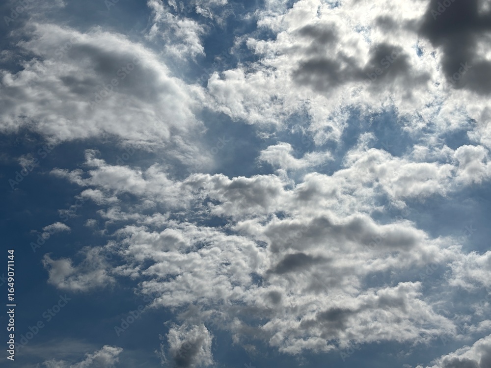 Obraz premium Bright blue sky with a smooth spread of white clouds, sunlight is seen spreading softly from behind the cloud layer in the middle, creating a calm and peaceful atmosphere.
