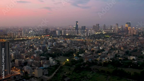 Aerial drone shot slow panning over gurgaon cityscape showing densely populated houses elevated roads, skyscrapers and water pools against the pink orange dusk sky