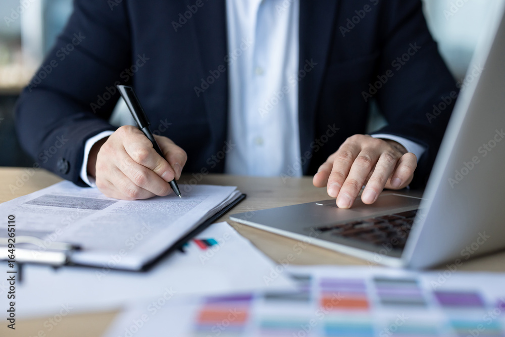 Close-up photo of the hands of a young man in a suit sitting at a desk in an office, typing on a laptop keyboard and writing a paper Close-up photo of the hands of a young man in a suit sitting at a desk in an office, typing on a laptop keyboard and writing a paper
