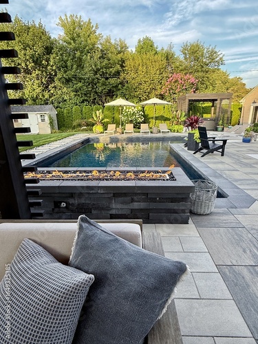 Beautiful, luxury, modern backyard lounge area. Umbrella, sofa, lounge chairs table poolside. Hotel resort feel. Seating and tanning sundeck by gray stone pavers. Fire pit and waterfall. High end. 