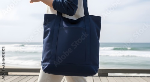 A person carries a large navy blue tote bag while walking on a wooden boardwalk near a beach with ocean waves.
