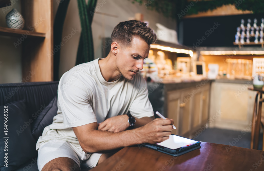 Fototapeta premium Freelancer drawing with stylus on tablet at café-style table, portraying deep creative focus, mobile work habits, and integration of digital tools into everyday professional life.