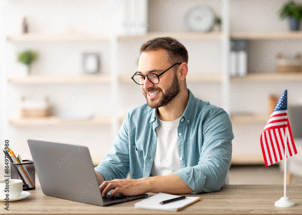 Happy young guy with flag of the US working at desk with laptop in home office. Millennial male student learning English online for foreign job, education or emigration to America Happy young guy with flag of the US working at desk with laptop in home office. Millennial male student learning English online for foreign job, education or emigration to America