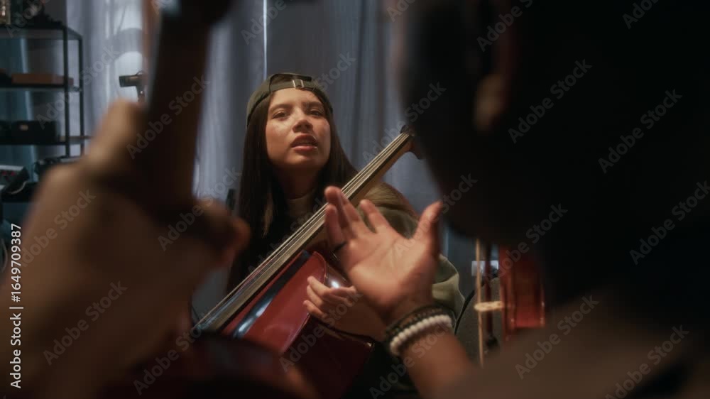 Young woman playing cello and discussing new composition with bandmates during music rehearsal in studio, over shoulder shot Young woman playing cello and discussing new composition with bandmates during music rehearsal in studio, over shoulder shot