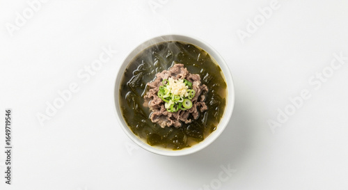 A steaming hot bowl of Korean beef and seaweed soup, miyeok-guk, garnished with scallions and garlic on a white background.