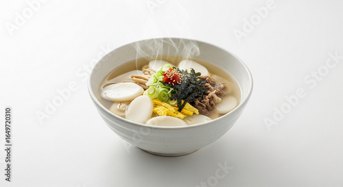 A steaming bowl of traditional Korean rice cake soup, Tteokguk, with beef, egg, and seaweed garnish on a white background.