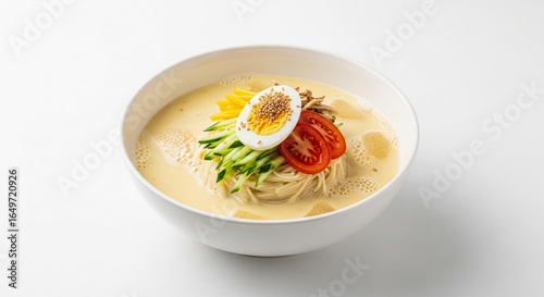 A white bowl of Korean cold soy milk noodle soup, Kongguksu, garnished with a boiled egg, cucumber, and tomato on a white background.