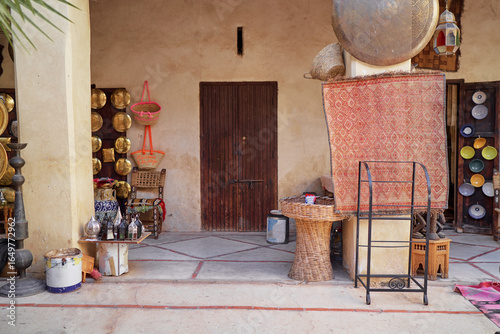 A traditional market stall displaying various metalware and woven baskets, with a rustic wooden door and a red patterned rug hanging on the wall. Travel by Morocco.
