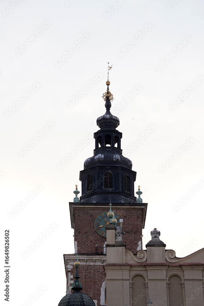 Obraz premium A tall brick clock tower with dark domed spire and golden ornaments rises above ornate facades, symbolizing timeless architecture and the silent passage of centuries.