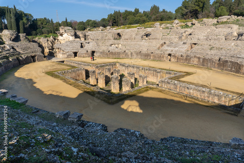 Ancient Roman Amphitheater itálica: A grand stone amphitheater stands proudly against a backdrop of verdant trees, the arena's walls weathered by time and history.