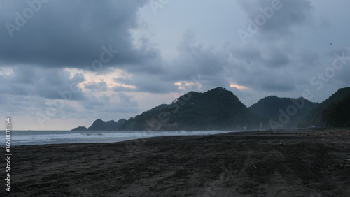 Dramatic seascape showing dark sand, cloudy skies, and misty hills along the shoreline during a moody, overcast day.	