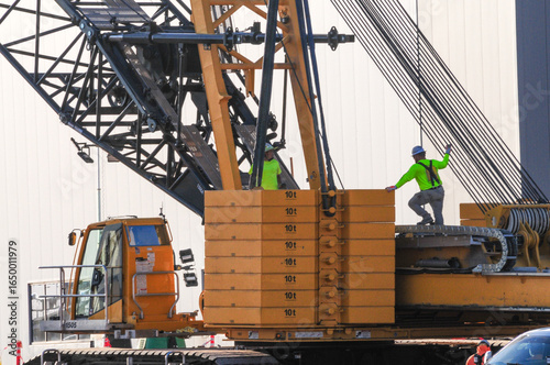 Construction workers secure rigging on a large crane at a stand-alone electric battery manufacturing plant under construction, with counterweights and lifting components visible on heavy equipment