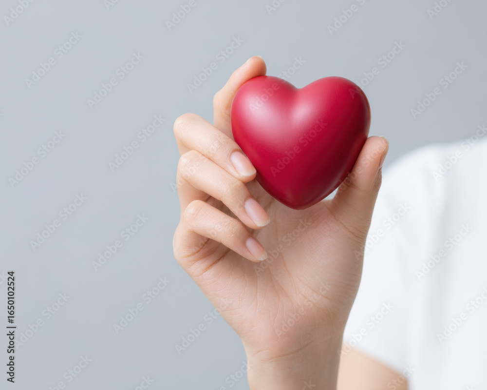 Obraz premium Close up of hand gently holding smooth red heart shape, symbolizing love and care with soft gray background