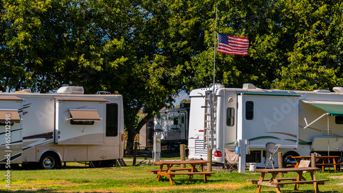 Several recreational vehicles are parked under a lush canopy of trees, creating a serene campground scene. An American flag flutters in the gentle breeze, while picnic tables invite visitors to relax.