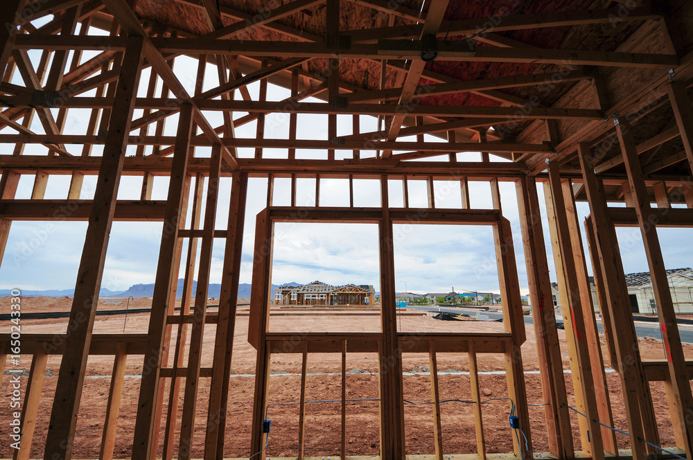 Obraz premium Wood framing of a residential home under construction shows interior wall studs, window openings, and roof trusses, with additional new houses visible across the future subdivision development site