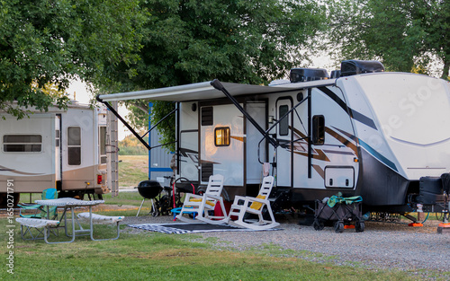 An inviting campsite features a modern RV with an extended awning, under which two white lounge chairs are positioned for relaxation. A picnic table and grill setup complete the cozy outdoor living 