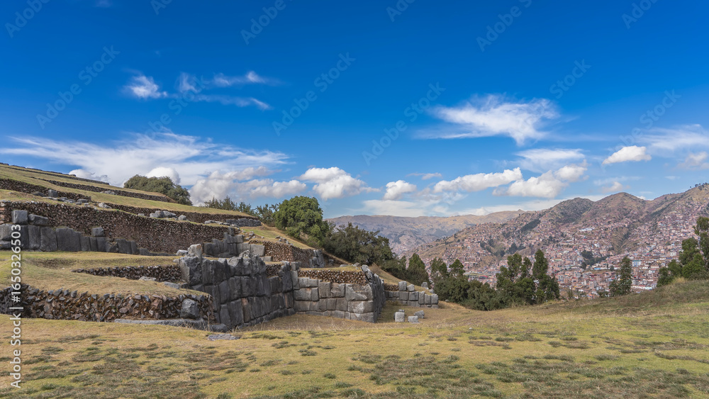 Obraz premium Ruins of the majestic ancient Inca fortress Saksaywaman. Terraces and tiers of walls made of polished large stones. Green grass on a meadow. In the distance, on a mountainside, the Cusco city