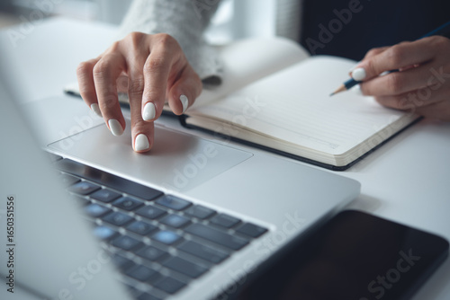 Business woman using laptop computer to search the information and taking note on notebook on office table. Student studying online, jotting down on notepad, e-learning, business planning concept
