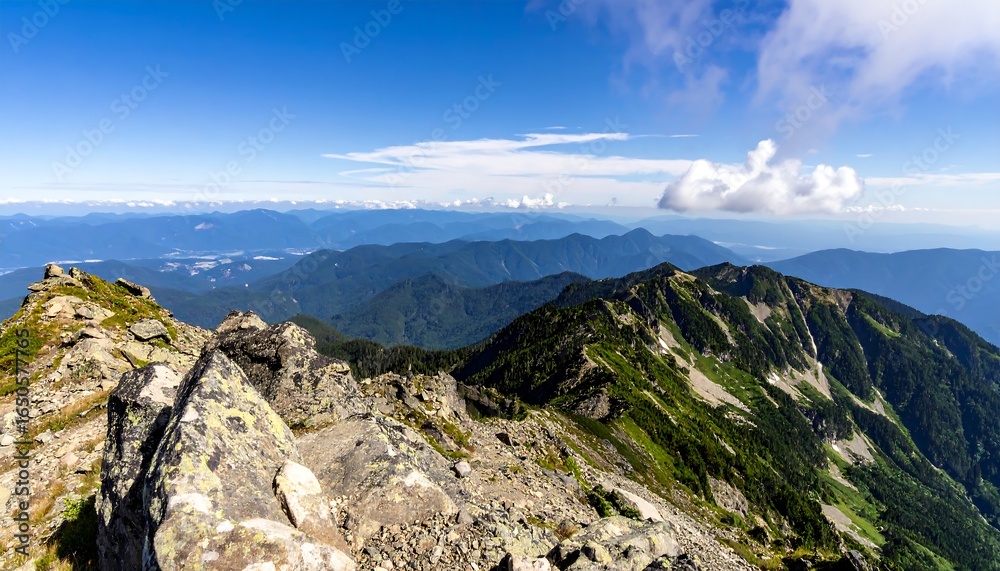 Fototapeta premium Panoramic view of a mountain range, showing rocky foreground and lush green slopes receding into distant blue mountains under a partly cloudy sky