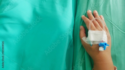 Close-up of a hand with an IV drip attached, lying on a hospital bed with a green sheet and patient wristband visible.