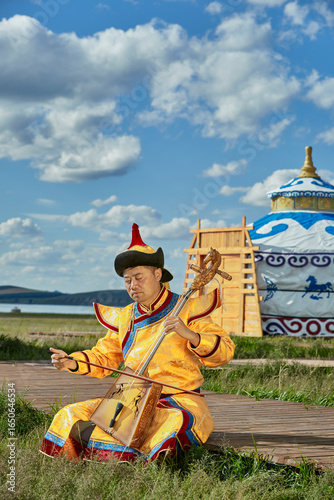 Obraz na plátně A man dressed in Mongolian attire playing the morin khuur (horsehead fiddle) in front of a yurt