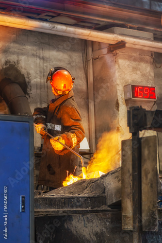 A man in a fireproof suit and helmet measures the temperature of hot metal in a metallurgical furnace with a thermometer and looks at the thermometer