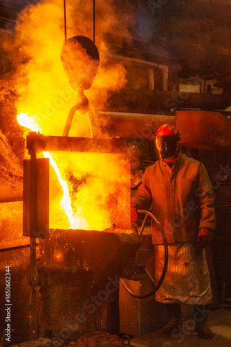A man in a fireproof suit and helmet turns over molten burning metal with fiery splashes from a ladle, which pours out in a fiery stream from a metallurgical furnace