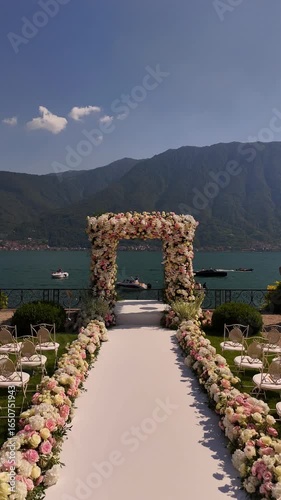 Aerial drone view of luxury destination wedding at Lake Como, Italy. White aisle, floral arch, and neat chairs set against breathtaking lake and mountain scenery